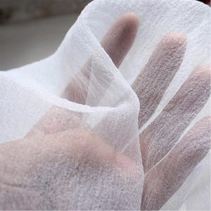 Op de afbeelding: Close-up van een doorschijnende witte stof, tegen het licht gehouden. De stof is fijn geweven, met een delicate, luchtige textuur. Licht gaat door het materiaal, waardoor de weefselstructuur wordt benadrukt en een zacht, etherisch effect ontstaat. De stof lijkt licht en vloeiend.