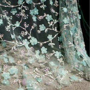 May include: Delicate, sheer fabric with a floral design. The fabric features light blue 3D flowers and embroidered vines with white and lavender accents. The material is layered, creating a textured effect. Ideal for crafting or special occasion garments.