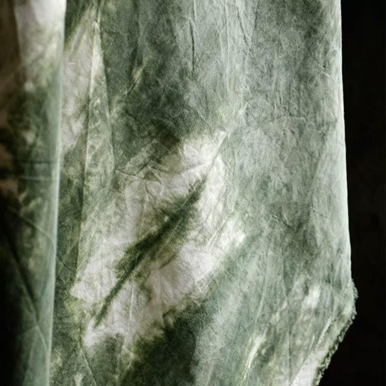 Puede incluir: Primer plano de una tela con un estampado tie-dye. La tela es principalmente blanca con manchas y vetas verdes y verde oscuro. La tela est&aacute; arrugada y tiene una textura suave. La tela probablemente se utiliza para ropa o decoraci&oacute;n del hogar.