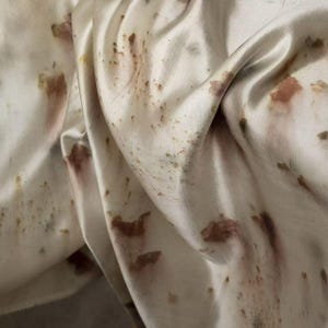 Op de afbeelding: Close-up van een crèmekleurige stof met bruine en groene organische patronen. De stof is gedrapeerd en toont plooien en texturen. De patronen lijken natuurlijk, mogelijk van een verf- of drukproces, waardoor een uniek, artistiek textiel ontstaat.