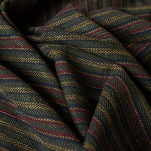May include: Close-up of a folded fabric with a herringbone pattern. The fabric features alternating stripes of dark green, navy blue, gold, and burgundy. The texture appears soft and the fabric is draped.