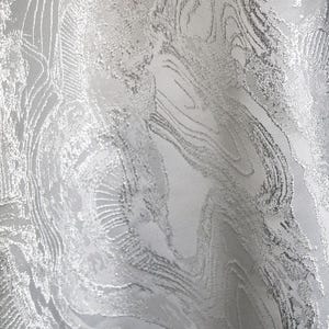 K&ouml;nnte beinhalten: Nahaufnahme eines silbernen Stoffes mit strukturiertem, marmoriertem Aussehen. Der Stoff hat ein wirbelndes Muster mit erhabenen, metallisch aussehenden Details. Das Design &auml;hnelt flie&szlig;endem Wasser oder der Maserung in Stein.