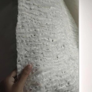 Op de afbeelding: Close-up van een getextureerde, gebroken witte stof. Het materiaal heeft een poreuze, bijna sponsachtige uitstraling met zichtbare gaten en een enigszins ongelijkmatig oppervlak. De stof wordt vastgehouden, waardoor de dikte en textuur zichtbaar worden.