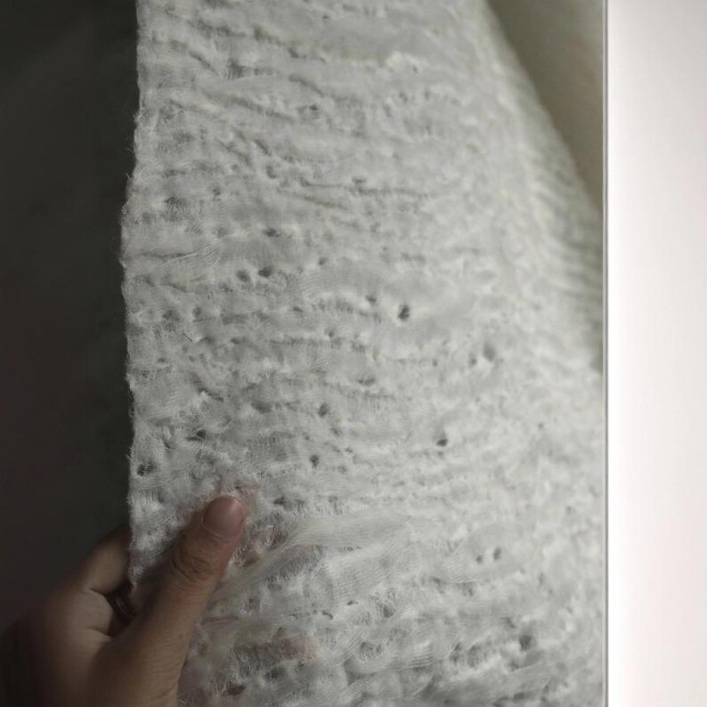 Op de afbeelding: Close-up van een getextureerde, gebroken witte stof. Het materiaal heeft een poreuze, bijna sponsachtige uitstraling met zichtbare gaten en een enigszins ongelijkmatig oppervlak. De stof wordt vastgehouden, waardoor de dikte en textuur zichtbaar worden.