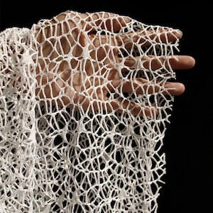 May include: Close-up of a white, open-weave fabric with a honeycomb-like pattern. The fabric is held up against a dark background, showcasing its delicate texture and design. The material appears to be soft and flexible.
