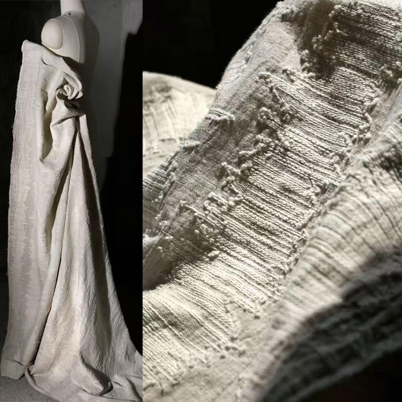 May include: A draped, off-white fabric with a textured surface is displayed on a mannequin. The fabric has a woven appearance with visible threads and raised, patterned details. The overall aesthetic is soft and natural, suggesting a focus on texture and organic materials.