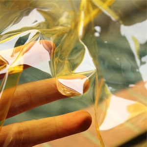May include: A flexible, transparent yellow plastic sheet with a smooth, glossy surface. The material is thin and pliable, showing light reflections and folds throughout its structure. The clear yellow tint allows for visibility through the layer.