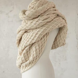 May include: A cream-colored, knitted infinity scarf with a textured, diamond-patterned design. The scarf is draped over a mannequin, showcasing its soft, cozy appearance. The neutral color and intricate knitwork suggest a handmade, winter accessory.