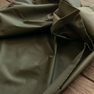 May include: Close-up of olive green fabric with a slight sheen. The fabric is folded and draped, creating shadows and highlights. The material appears smooth and possibly water-resistant, suitable for apparel or home decor projects.