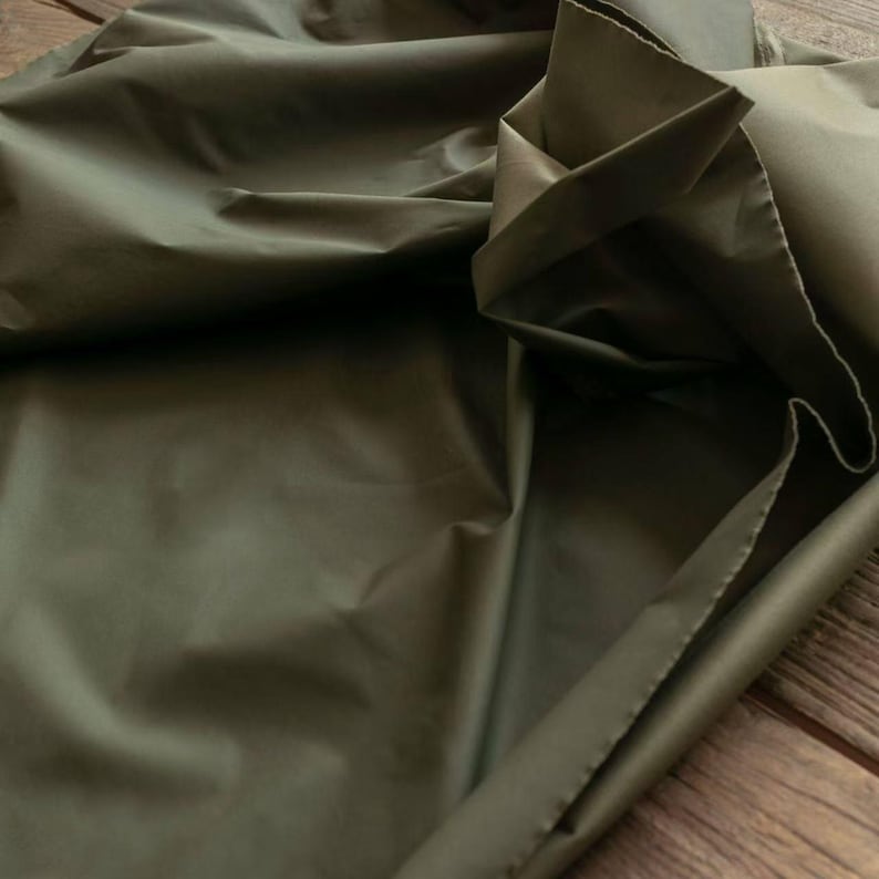 May include: Close-up of olive green fabric with a slight sheen. The fabric is folded and draped, creating shadows and highlights. The material appears smooth and possibly water-resistant, suitable for apparel or home decor projects.