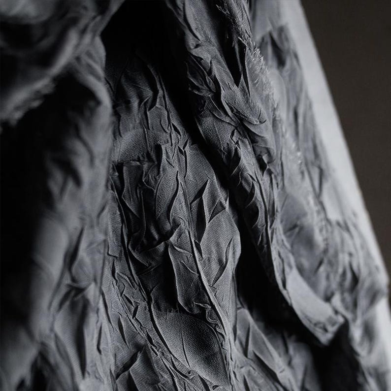 May include: Close-up of a dark gray fabric with a textured, leaf-like pattern. The fabric appears to be draped, with folds and shadows creating depth. The material has a matte finish, and the overall effect is elegant and sophisticated.