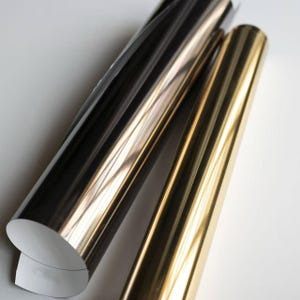 Könnte beinhalten: Drei Rollen Metallic-Vinyl in Schwarz, Silber und Gold. Die Rollen sind eng gewickelt, wobei die Kanten des Vinyls das Licht reflektieren. Die Rollen befinden sich auf einem weißen Hintergrund, wodurch ein Kontrast entsteht, der den metallischen Glanz hervorhebt.