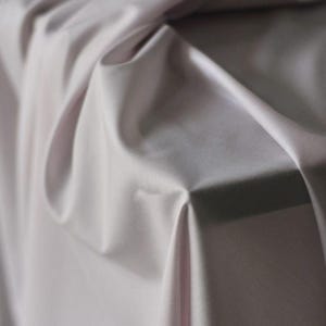 May include: Close-up of a draped fabric in a soft, muted lavender hue. The material has a smooth, silky texture with subtle folds and shadows, creating depth and dimension. The fabric appears to be lightweight and flowing, suitable for apparel or home decor.
