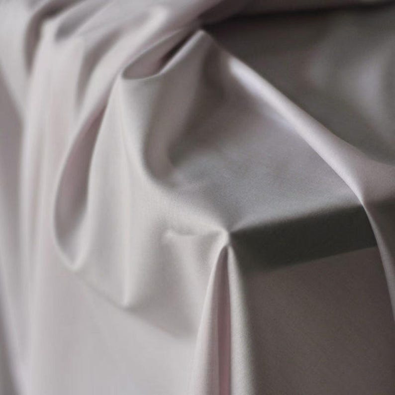 May include: Close-up of a draped fabric in a soft, muted lavender hue. The material has a smooth, silky texture with subtle folds and shadows, creating depth and dimension. The fabric appears to be lightweight and flowing, suitable for apparel or home decor.