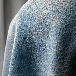 Op de afbeelding: Close-up van een textiel met een netwerk van witte lijnen op een blauwe achtergrond. De stof lijkt gedrapeerd, met schaduwen die diepte toevoegen aan het ontwerp. Het algehele effect is een gedetailleerd, abstract patroon.