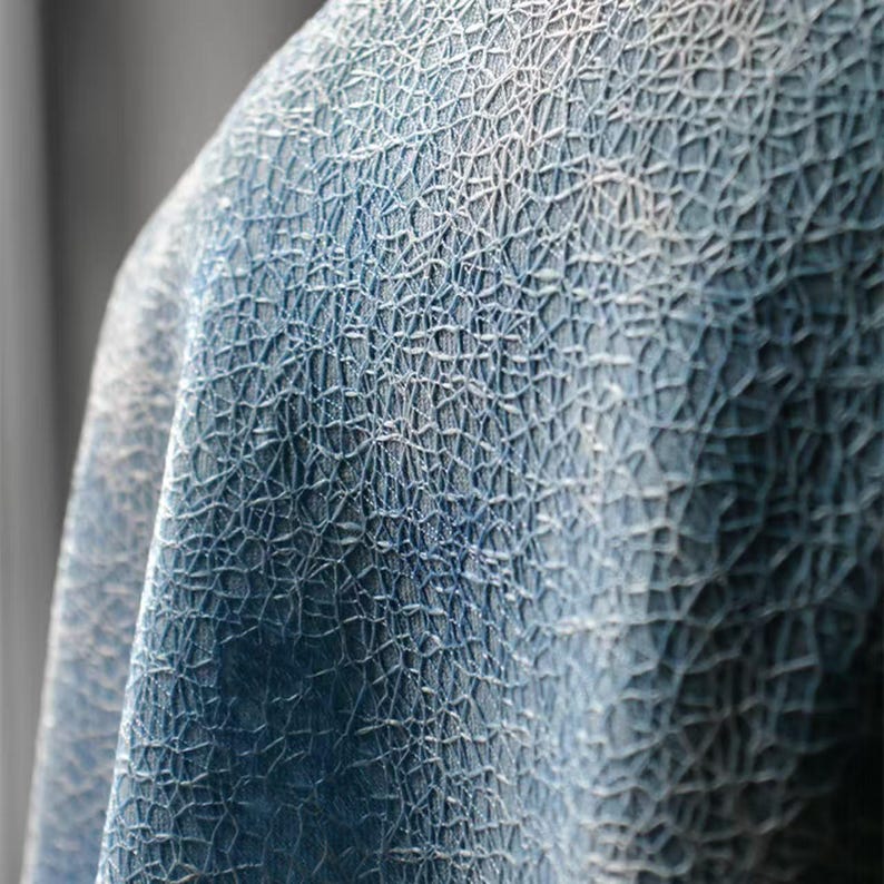 Op de afbeelding: Close-up van een textiel met een netwerk van witte lijnen op een blauwe achtergrond. De stof lijkt gedrapeerd, met schaduwen die diepte toevoegen aan het ontwerp. Het algehele effect is een gedetailleerd, abstract patroon.