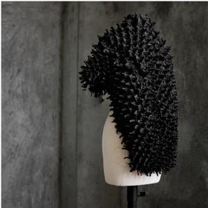 May include: A black, textured head covering, possibly a wig or hat, displayed on a mannequin head. The head covering has a dense, spiky texture and is set against a gray background. The overall aesthetic is modern and artistic.