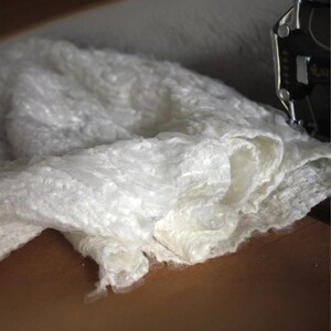 Op de afbeelding: Close-up van een witte, getextureerde stof met een zacht, gerimpeld uiterlijk. De stof is gelaagd, waardoor diepte en dimensie ontstaat. Het materiaal lijkt delicaat en kan worden gebruikt voor kleding of woondecoratie.