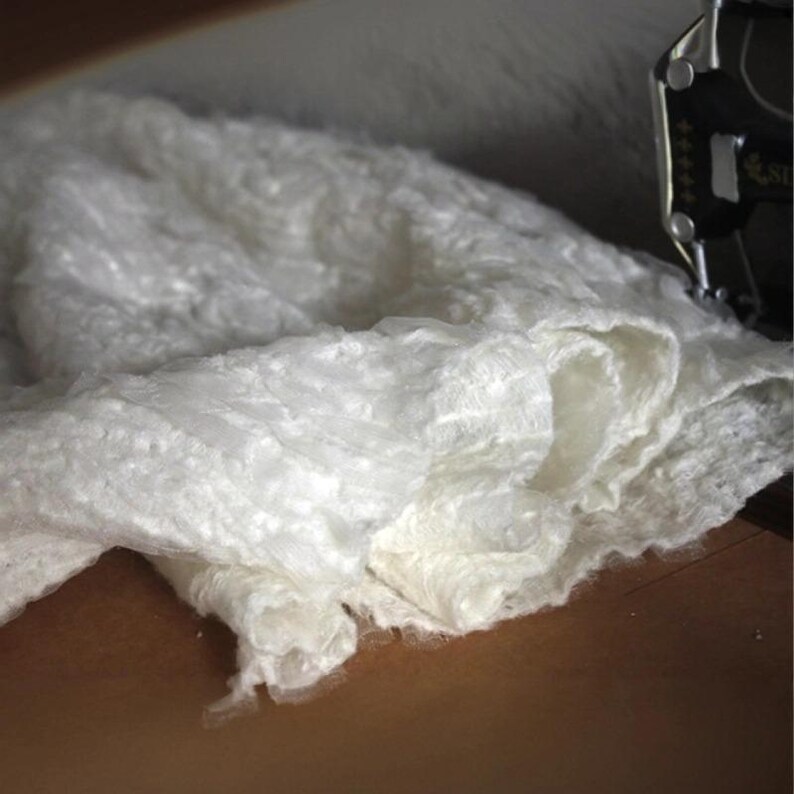 Op de afbeelding: Close-up van een witte, getextureerde stof met een zacht, gerimpeld uiterlijk. De stof is gelaagd, waardoor diepte en dimensie ontstaat. Het materiaal lijkt delicaat en kan worden gebruikt voor kleding of woondecoratie.