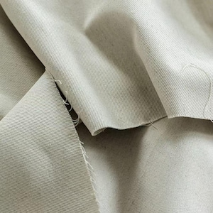 Könnte beinhalten: Nahaufnahme eines hellgrauen Stoffes mit sichtbarer Textur und Falten. Der Stoff scheint eine Naturfaser zu sein, möglicherweise Leinen oder Baumwolle, mit einer leicht rauen Oberfläche. Die Kanten sind unfertig und zeigen den rohen Schnitt des Materials.