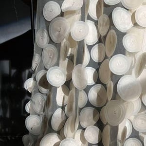 May include: A sheer, off-white fabric curtain adorned with numerous, layered, circular, petal-like embellishments. The circular shapes are of varying sizes and are attached to the sheer fabric, creating a textured, dimensional effect. The curtain hangs vertically.