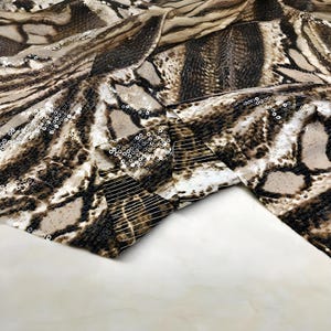 May include: Close-up of a fabric with a snakeskin pattern in shades of brown, beige, and black. The fabric is embellished with small, shiny sequins. The material is folded, revealing the texture and design.