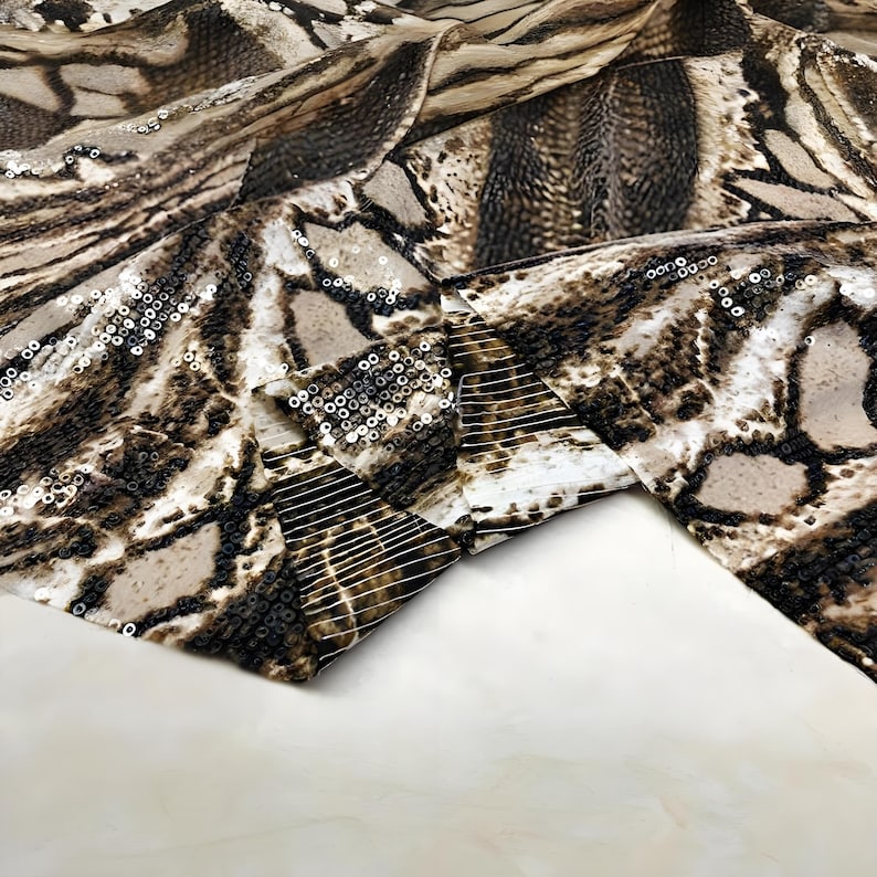 May include: Close-up of a fabric with a snakeskin pattern in shades of brown, beige, and black. The fabric is embellished with small, shiny sequins. The material is folded, revealing the texture and design.