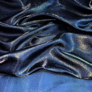 Puede incluir: Primer plano de una tela azul oscuro con un brillo brillante e iridiscente. La tela tiene una apariencia fluida y drapeada con reflejos dorados y verde azulado. La textura parece suave y lujosa, con pliegues y sombras sutiles.