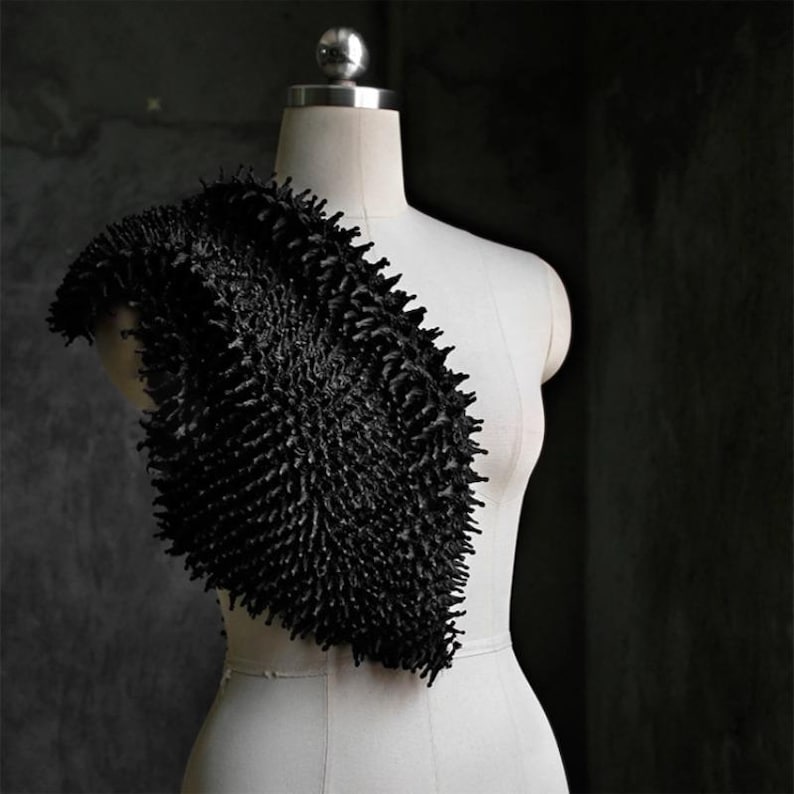 May include: A black, textured shoulder wrap displayed on a white dress form. The wrap has a dense, spiky appearance, creating a dramatic and avant-garde aesthetic. The background is a dark, textured wall, enhancing the contrast.