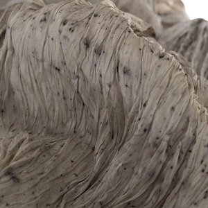 Könnte beinhalten: Nahaufnahme eines strukturierten Stoffes mit fließendem, drapiertem Aussehen. Der Stoff ist überwiegend hell und neutral gefärbt, mit dezenten dunklen Sprenkeln. Die Falten und Knitter erzeugen ein Gefühl von Tiefe und Bewegung.