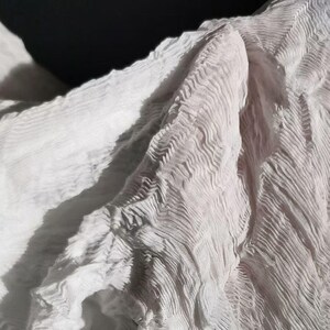 May include: Close-up of a white, textured fabric with a rippled, wave-like pattern. The fabric is draped, creating shadows and highlights that emphasize the texture. The material appears soft and lightweight, with a subtle sheen.