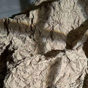 Może przedstawiać: Zbliżenie pogniecionej, teksturowanej powierzchni papieru. Papier ma stonowany brązowy kolor z obszarami cieni i świateł, tworząc poczucie głębi i wymiaru. Ogólny efekt jest abstrakcyjny i dotykowy.