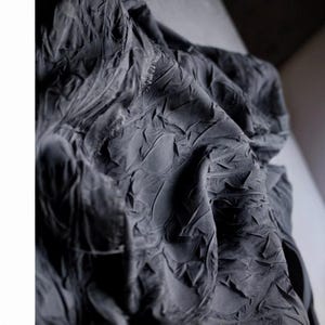 May include: Close-up of a dark gray fabric with a textured, wrinkled surface. The fabric appears to be draped, with folds and creases creating shadows and highlights. The material has a matte finish.