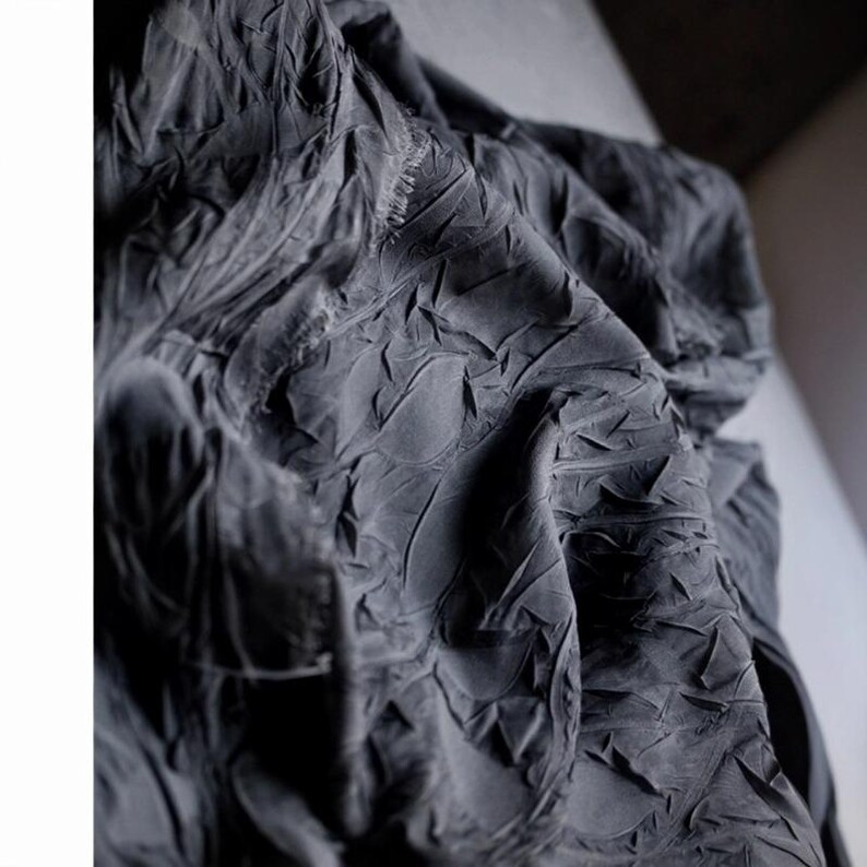 May include: Close-up of a dark gray fabric with a textured, wrinkled surface. The fabric appears to be draped, with folds and creases creating shadows and highlights. The material has a matte finish.