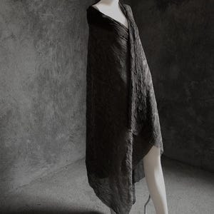 May include: A dark gray, textured shawl draped over a mannequin. The fabric has a crinkled appearance, creating a unique visual texture. The shawl is long and flowing, with a draped design. The background is a solid gray.