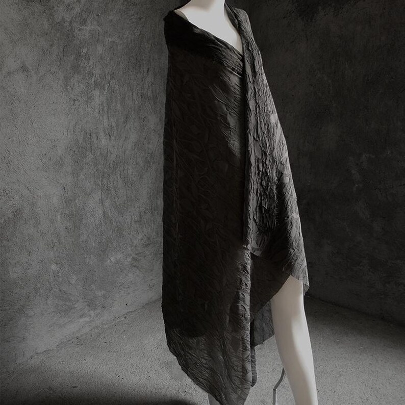 May include: A dark gray, textured shawl draped over a mannequin. The fabric has a crinkled appearance, creating a unique visual texture. The shawl is long and flowing, with a draped design. The background is a solid gray.