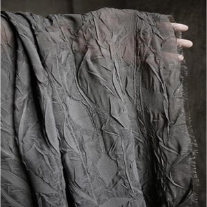 May include: A close-up of a dark gray fabric with a textured, leaf-like pattern. The fabric is draped, revealing its delicate, semi-sheer quality. The edges are frayed, adding a touch of rustic charm to the textile.