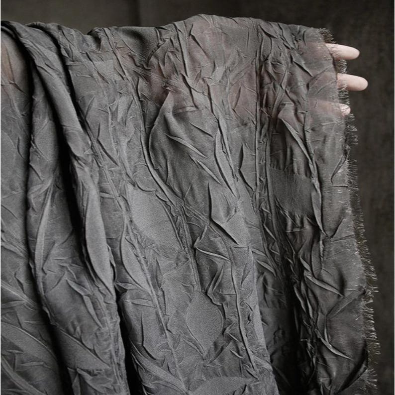 May include: A close-up of a dark gray fabric with a textured, leaf-like pattern. The fabric is draped, revealing its delicate, semi-sheer quality. The edges are frayed, adding a touch of rustic charm to the textile.