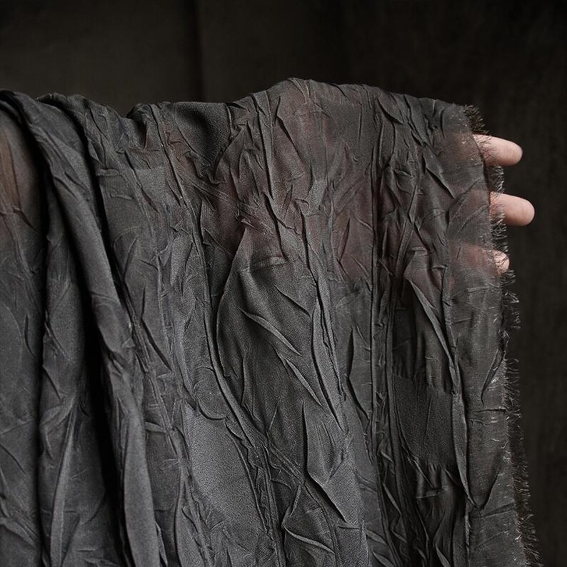 May include: Dark gray fabric with a crinkled texture, held up to show its draping. The fabric has a slightly sheer quality, with visible folds and a frayed edge. The overall appearance is soft and flowing, with a matte finish.