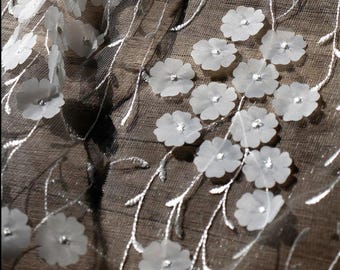 May include: Close-up of a sheer fabric with white, three-dimensional flower appliqués. The flowers have a small, sparkling gem in the center. Silver-colored embroidered vines with leaves are also visible.