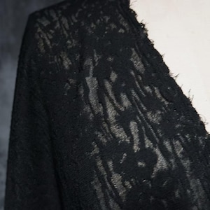 May include: A black garment featuring a textured, semi-sheer lace fabric with a floral pattern. The piece has a V-neckline with a raw, frayed edge detail along the collar, showcasing a delicate, intricate weave throughout the material.