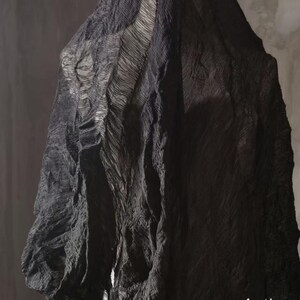 May include: A draped, textured fabric in shades of black and gray. The material appears to have a layered, rippled design, creating a sense of depth and movement. The fabric is likely used for clothing or decorative purposes.