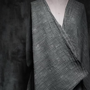 Può includere: Un capo grigio scuro aperto sul davanti con scollo a V, drappeggiato su un manichino. Il tessuto presenta un motivo distintivo testurizzato, stropicciato e a righe, creando un aspetto stratificato con linee di trama orizzontali e verticali.