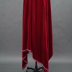 May include: A rich, deep red velvet fabric, draped over a black stand. The fabric has a soft texture and is trimmed with a white stitched edge. The folds create shadows, highlighting its luxurious appearance. The overall theme is elegant and theatrical.