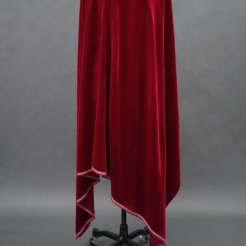 May include: A rich, deep red velvet fabric, draped over a black stand. The fabric has a soft texture and is trimmed with a white stitched edge. The folds create shadows, highlighting its luxurious appearance. The overall theme is elegant and theatrical.