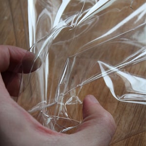 May include: A sheet of clear, flexible, and transparent plastic material with a smooth, glossy surface. The material is thin and pliable, showing multiple folds, creases, and light reflections across its surface as it is being manipulated.