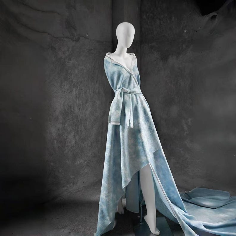 Op de afbeelding: Lichtblauwe jurk met een strik in de taille en een vloeiende, asymmetrische rok, gepresenteerd op een mannequin. De jurk heeft een capuchon en een subtiel, wolkenachtig patroon. De achtergrond is donkergrijs.