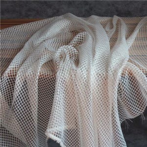 Puede incluir: Una tela blanca transparente que presenta un patrón de cuadrícula consistente de líneas que se cruzan. El material está drapeado en pliegues suaves, creando una apariencia texturizada con espacios visibles entre los hilos tejidos en toda la pieza.