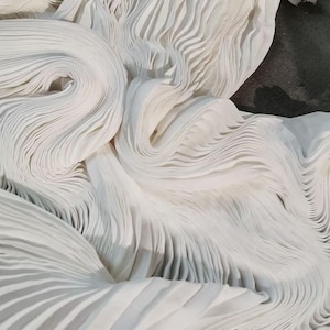 Op de afbeelding: Close-up van witte geplooide stof, met zachte, vloeiende plooien. De stof is gerangschikt in wervelende patronen, waardoor een gevoel van beweging en textuur ontstaat. Het licht reflecteert op de stof en benadrukt de delicate plooien. Het algehele effect is elegant en etherisch.