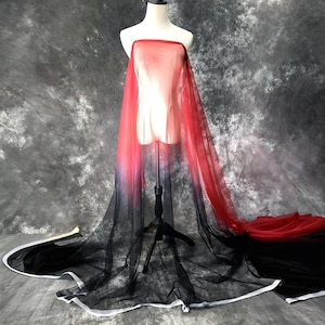 May include: A sheer fabric draped over a mannequin, transitioning from red at the top to black at the bottom. The fabric is lightweight and flowing, with a white trim along the edges of the black portion. The background is a mottled gray.